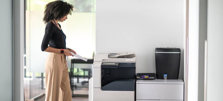 A stylish professional stands over an all-in-one copier, printer and fax machine in an office setting waiting for outputs, signifying Managed Print Services.