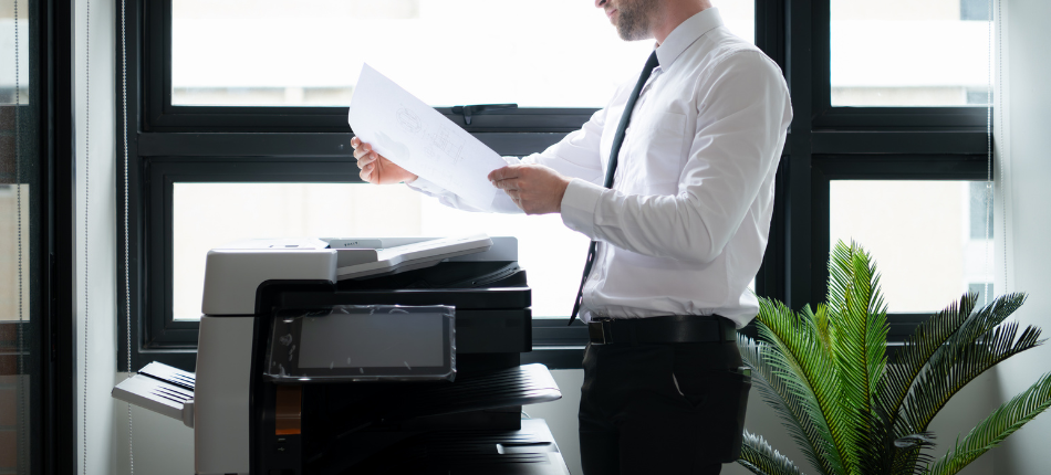 A professional stands in front of an office copier examining the output with a questioning expression.
