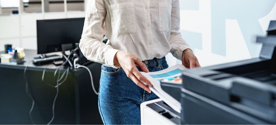 A person in a modern office setting is retrieving printed documents from a professional business printer, a common scene in workplaces that utilize a business printer lease to manage their document production needs efficiently.