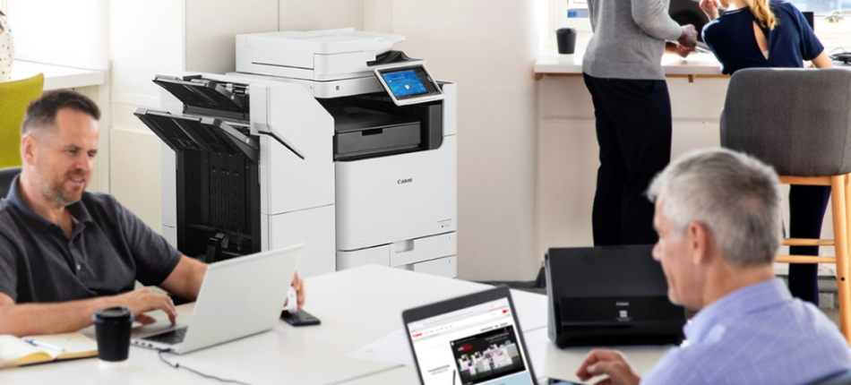 Office environment with employees working on laptops and a Canon laser printer in the background. Two employees are seated at a table, while another two are standing by a window. The laser printer is prominently displayed, highlighting its use in a busy office setting.