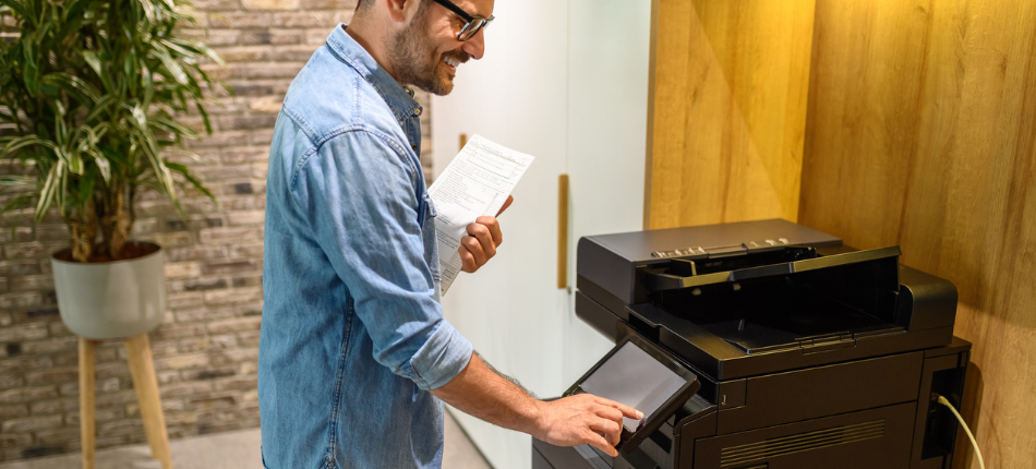 A man smiling while using a business multifunction printer in an office environment, holding a document and interacting with the touchscreen interface of the machine.