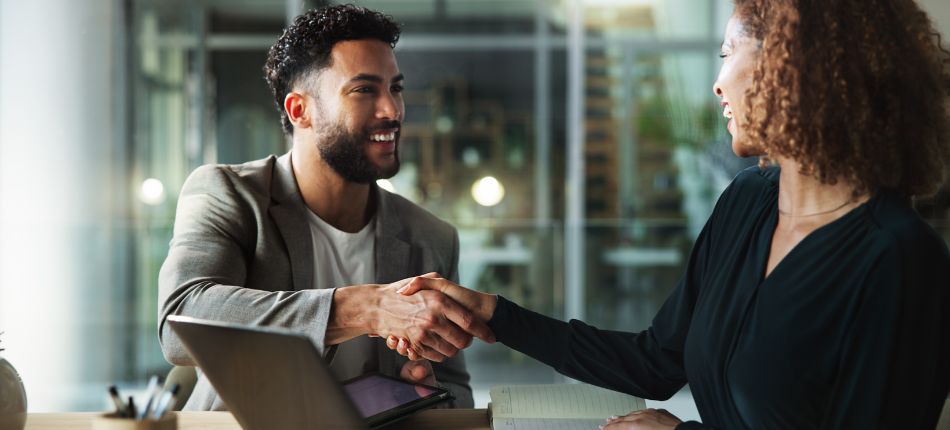 Two young professional shake hands while one holds a laptop at a desk signifying a printer lease agreement.